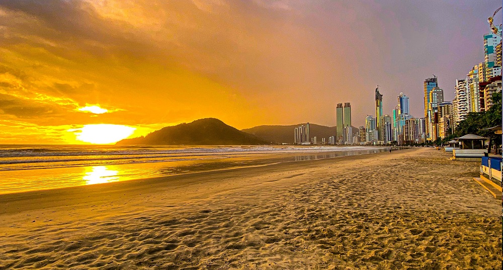 por do sol na praia, mostrando a areia, o céu amarelado refletindo na água do mar e os prédios de balneário camboriú ao fundo